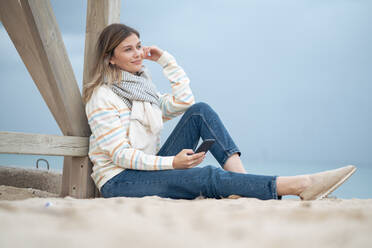 Nachdenkliche junge Frau sitzt mit Smartphone am Strand - JOSEF15448