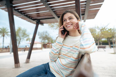 Lächelnde schöne Frau, die auf einer Bank sitzt und über ihr Mobiltelefon spricht - JOSEF15454
