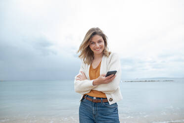 Glückliche schöne junge Frau mit Smartphone am Strand stehend - JOSEF15457
