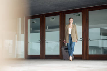 Smiling businesswoman walking with luggage in front of door - JOSEF15459
