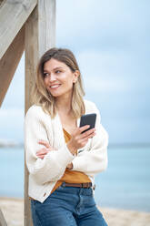 Lächelnde schöne junge Frau mit Smartphone an Holz gelehnt am Strand - JOSEF15469