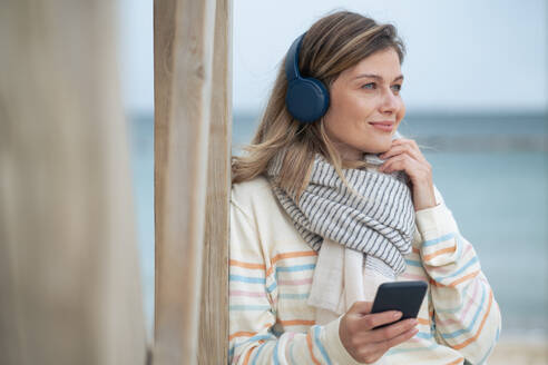 Lächelnde schöne junge Frau hält Smartphone und hört Musik am Strand - JOSEF15476