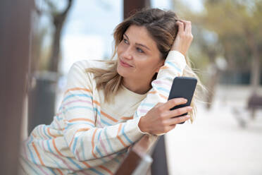 Thoughtful young blond woman sitting on bench with smart phone - JOSEF15483