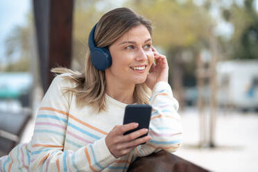 Lächelnde junge Frau, die ein Smartphone benutzt und Musik hört, auf einer Bank sitzend - JOSEF15485