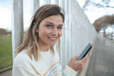 Lächelnde blonde Frau mit Smartphone am Zaun lehnend - JOSEF15494