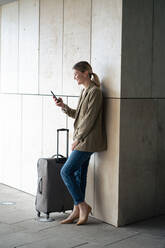 Smiling businesswoman using smart phone by luggage leaning on wall in tunnel - JOSEF15496