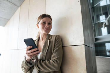Lächelnde Geschäftsfrau mit Smartphone vor einer Wand - JOSEF15497