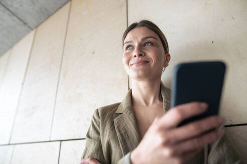 Lächelnde Geschäftsfrau mit Mobiltelefon vor einer Wand - JOSEF15498