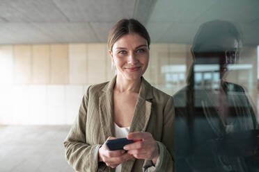 Smiling businesswoman with smart phone leaning on glass wall - JOSEF15505