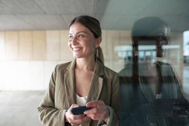 Happy businesswoman with smart phone leaning on glass wall - JOSEF15506