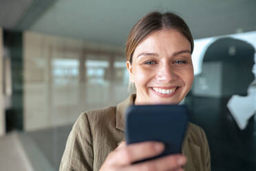 Lächelnde Geschäftsfrau, die ein Smartphone benutzt und sich an eine Glaswand lehnt - JOSEF15507