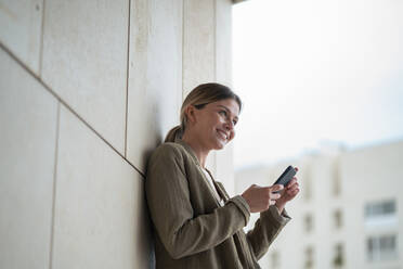 Smiling businesswoman with mobile phone in front of wall - JOSEF15508