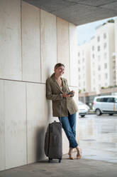 Smiling businesswoman leaning by luggage on wall - JOSEF15510