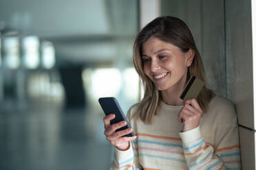 Lächelnde junge Frau mit Smartphone und Kreditkarte an der Wand lehnend - JOSEF15515