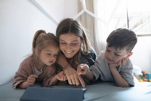 Lächelnde junge Mutter mit Kindern, die im Schlafzimmer zu Hause einen Tablet-PC benutzen - JOSEF15518