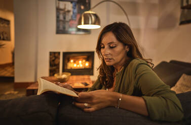 Side view of positive mature Hispanic female sitting on couch and reading book in weekend evening in living room with fireplace - ADSF41946