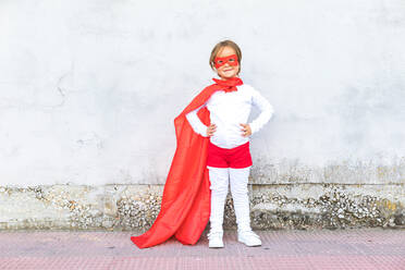 Full body of cute little girl in superhero costume and red cape standing with hands on waist against concrete wall - ADSF41955