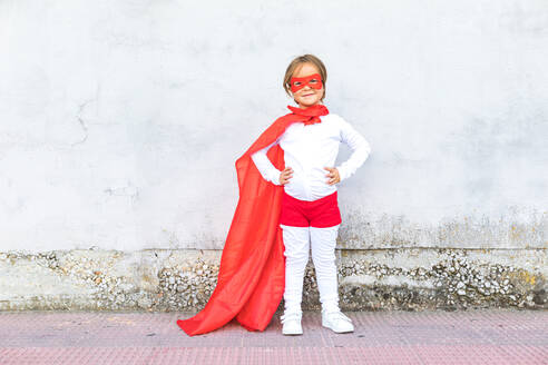 Ganzkörper des niedlichen kleinen Mädchens in Superheldenkostüm und rotem Umhang stehend mit Händen auf der Taille gegen Betonwand - ADSF41955