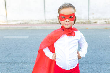Full body of cute little girl in superhero costume and red cape standing with hands on waist against concrete wall - ADSF41956