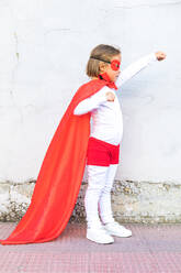 Full body side view of little girl in superhero costume and mask standing with raised arm and looking away against wall - ADSF41957