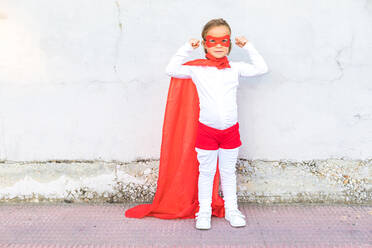 Full body of adorable little girl in superhero costume and mask flexing arm muscles looking at camera while standing against wall - ADSF41958