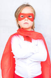 Cute little girl in superhero costume and red cape standing with crossed arms against concrete wall - ADSF41959