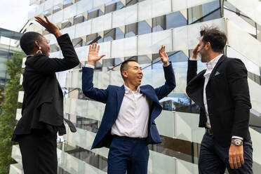 Happy multiracial coworkers in formal outfits standing against urban glass building and giving high five to each other while celebrating successful deal - ADSF41979