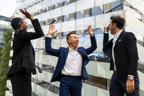 Glückliche multirassische Mitarbeiter in formellen Outfits, die vor einem städtischen Glasgebäude stehen und sich gegenseitig 