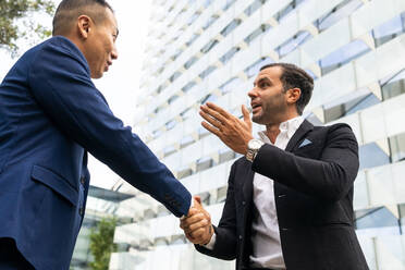 Low angle of positive multiracial businessmen in formal suits standing on street near modern multistory building and talking while shaking hands - ADSF41982