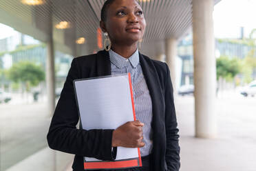 Thoughtful African American businesswoman with short hair in formal suit and earrings standing under entrance canopy of modern building and holding documents while looking away - ADSF41988