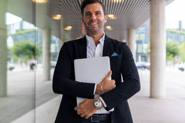 Positive caucasian male entrepreneur in formal suit standing under entrance canopy near glass wall of modern building and holding laptop while looking at camera - ADSF41989