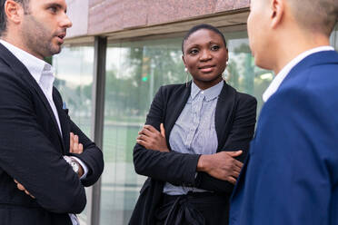 Positive afroamerikanische Frau in formellem Anzug, die mit verschränkten Armen an einer Glaswand steht und mit männlichen Kollegen eine Geschäftsstrategie bespricht - ADSF41994