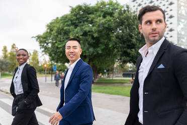 Side view of cheerful diverse coworkers in formal clothes walking along pedestrian crossing and chatting on street - ADSF41995