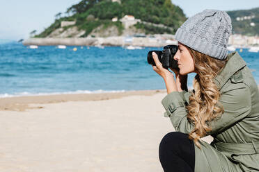 Side view of female photographer in knitted hat and trench coat sitting on haunches and shooting waving sea on sandy beach - ADSF42044