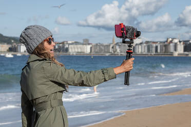 Side view of young woman with long hair in casual trench coat standing on sandy beach near waving sea and recording vlog via professional camera with microphone - ADSF42046