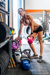 Fit smiling female athlete in sportswear looking at camera while lifting heavy kettlebell during workout at gym - ADSF42053