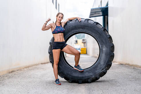 Positiver weiblicher Ganzkörpersportler, der in die Kamera schaut und seine Armmuskeln zeigt, während er auf einem riesigen Reifen während eines Funktionstrainings auf der Straße lernt - ADSF42060