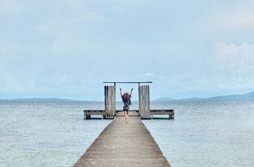 Back view of distant female walking on wooden shabby pier with gates and raising arms up enjoying freedom on sea - ADSF42094