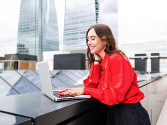 Happy woman in stylish red shirt touching neck and smiling while browsing data on netbook during work on remote project on city street - ADSF42110