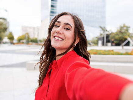 Optimistische junge Brünette in stilvollem rotem Hemd mit langen braunen Haaren schaut mit einem Lächeln in die Kamera, während sie ein Selfie auf der Straße macht - ADSF42111