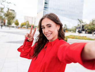 Optimistic young brunette in stylish red shirt with long brown hair gesturing V sign and looking at camera with smile while taking selfie on city street - ADSF42112