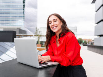 Positive female manager in stylish clothes with long brown hair looking at camera with smile and typing on netbook keyboard while working on remote project on street of modern city - ADSF42113