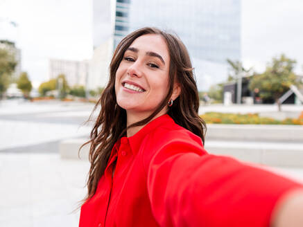Optimistische junge Brünette in stilvollem rotem Hemd mit langen braunen Haaren schaut mit einem Lächeln in die Kamera, während sie ein Selfie auf der Straße macht - ADSF42114