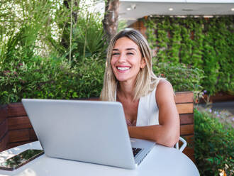 Glückliche blonde Frau, die lächelt und auf ihrem Netbook surft, während sie am Tisch sitzt und in die Kamera schaut und aus der Ferne auf der Terrasse arbeitet - ADSF42116