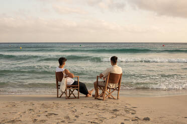 Back view full body of unrecognizable male friends in summer clothes sitting on fishing chairs and admiring picturesque wavy endless sea - ADSF42126