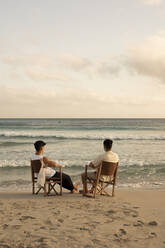 Back view full body of unrecognizable male friends in summer clothes sitting on fishing chairs and admiring picturesque wavy endless sea - ADSF42127