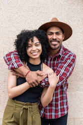 Fröhlicher kolumbianischer Mann in kariertem Hemd und Hut lächelt und umarmt seine Freundin von hinten, während er in der Nähe einer beigen Wand auf einer Straße steht - ADSF42133