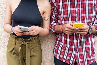 Cropped unrecognizable couple in casual clothes browsing on smartphones while standing on city street wall in weekend together - ADSF42134