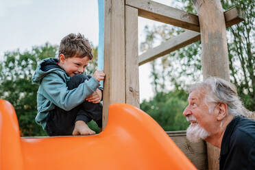 Fröhlicher barfüßiger Enkel lächelt und klopft auf Holz, während er in der Nähe der Rutsche sitzt und mit seinem Opa auf dem Spielplatz spielt - ADSF42135