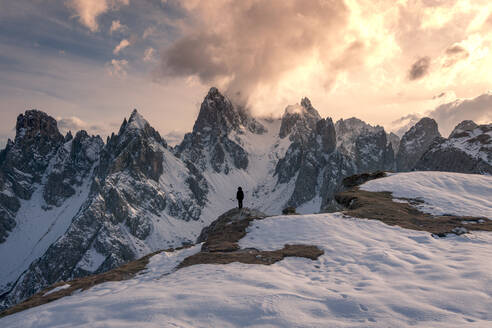 Rückenansicht eines nicht erkennbaren Touristen in Oberbekleidung, der auf Schnee steht und die Gipfel der Drei Zinnen gegen den wolkenverhangenen Himmel bei Sonnenuntergang in den Dolomiten, Italien, betrachtet - ADSF42143